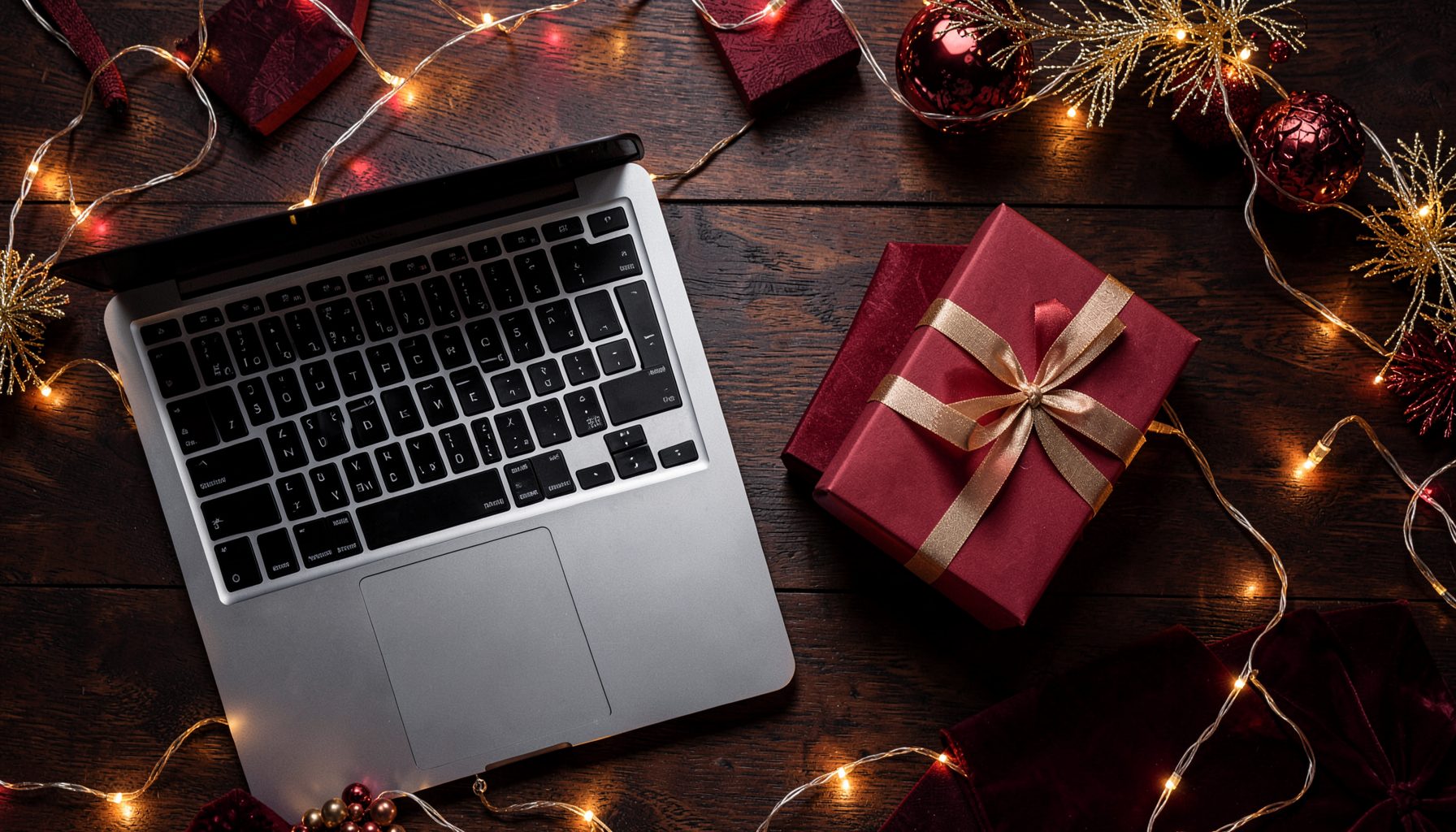 A festive holiday-themed flat lay with deep red and gold tones, featuring a sleek laptop beside a wrapped smartphone gift box, accented by warm fairy lights on a dark wooden surface.