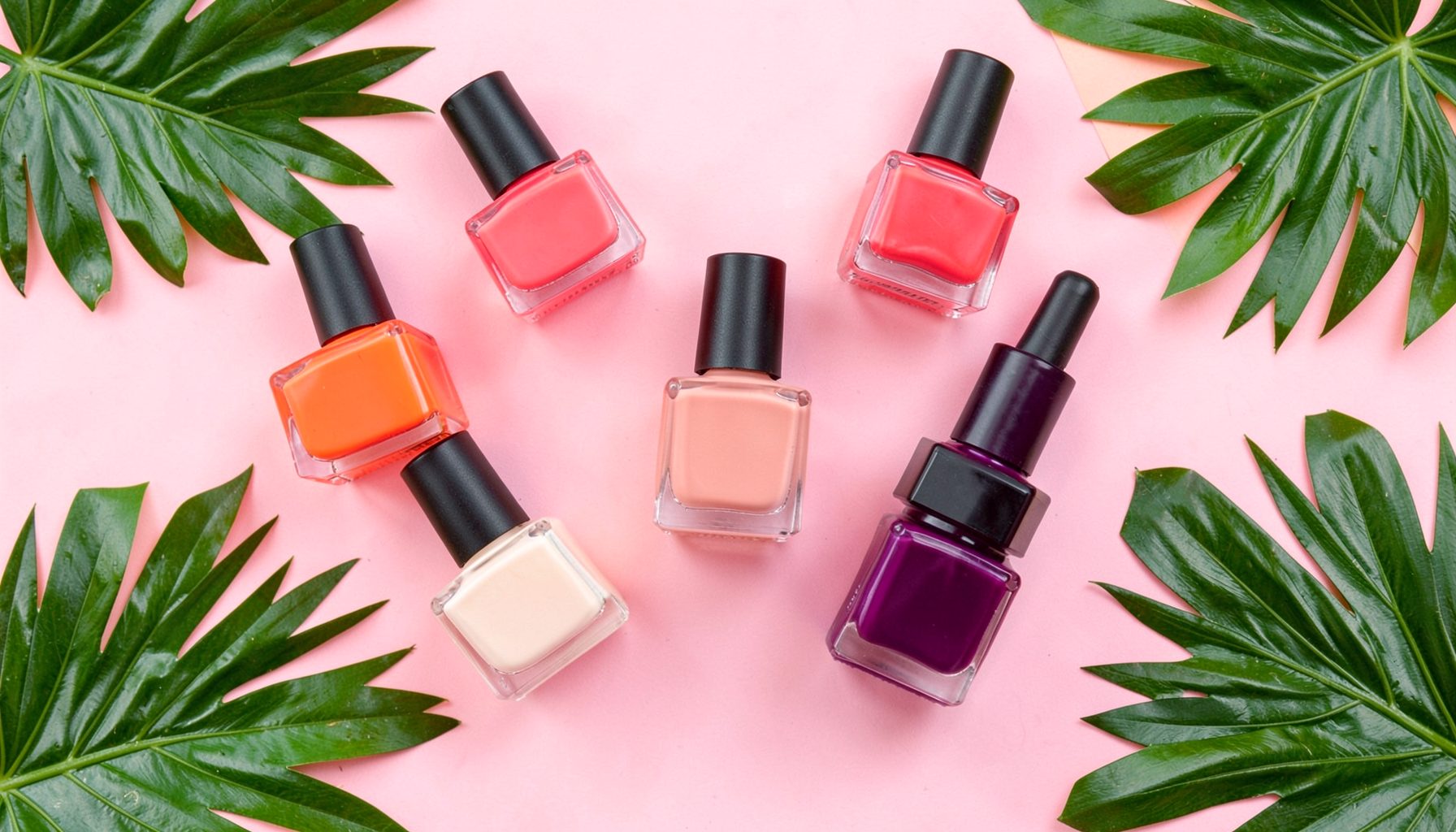 A vibrant flat-lay of colorful nail polish bottles in coral, blush, and berry tones with tropical leaves, bright and playful